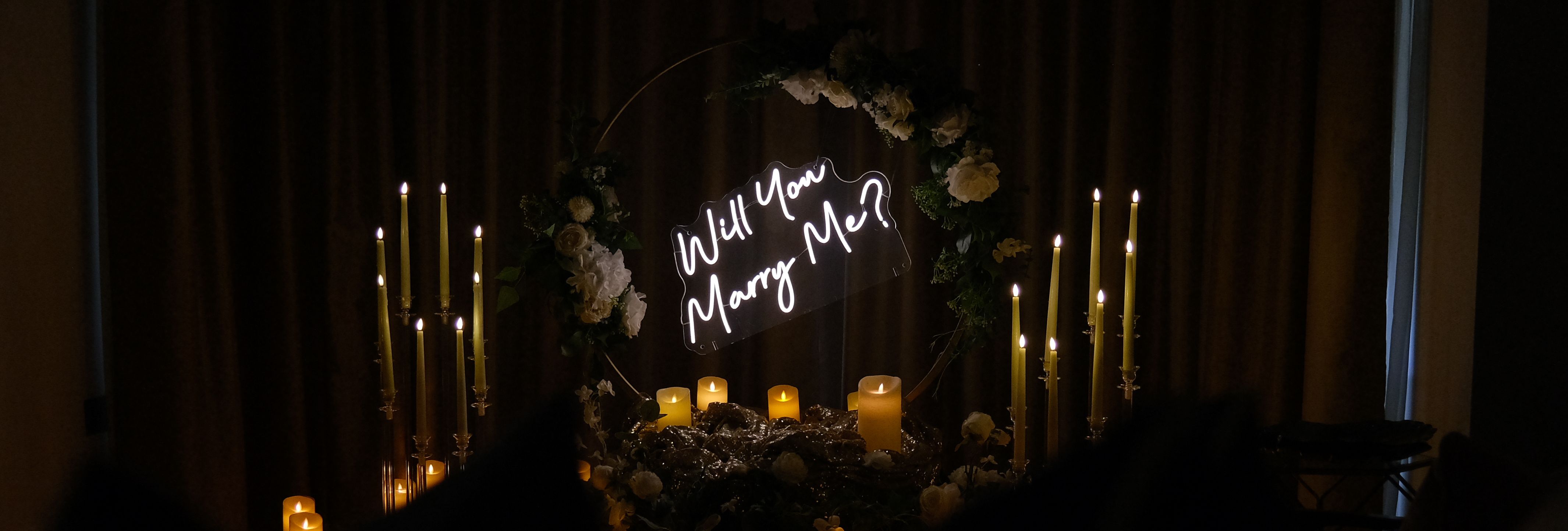 Romantic candlelit proposal set-up with illuminated 'Will You Marry Me?' sign and floral arch