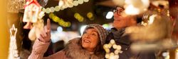 A smiling couple enjoys a Christmas market, admiring festive decorations. The scene is illuminated by warm string lights, with holiday ornaments and trinkets surrounding them.