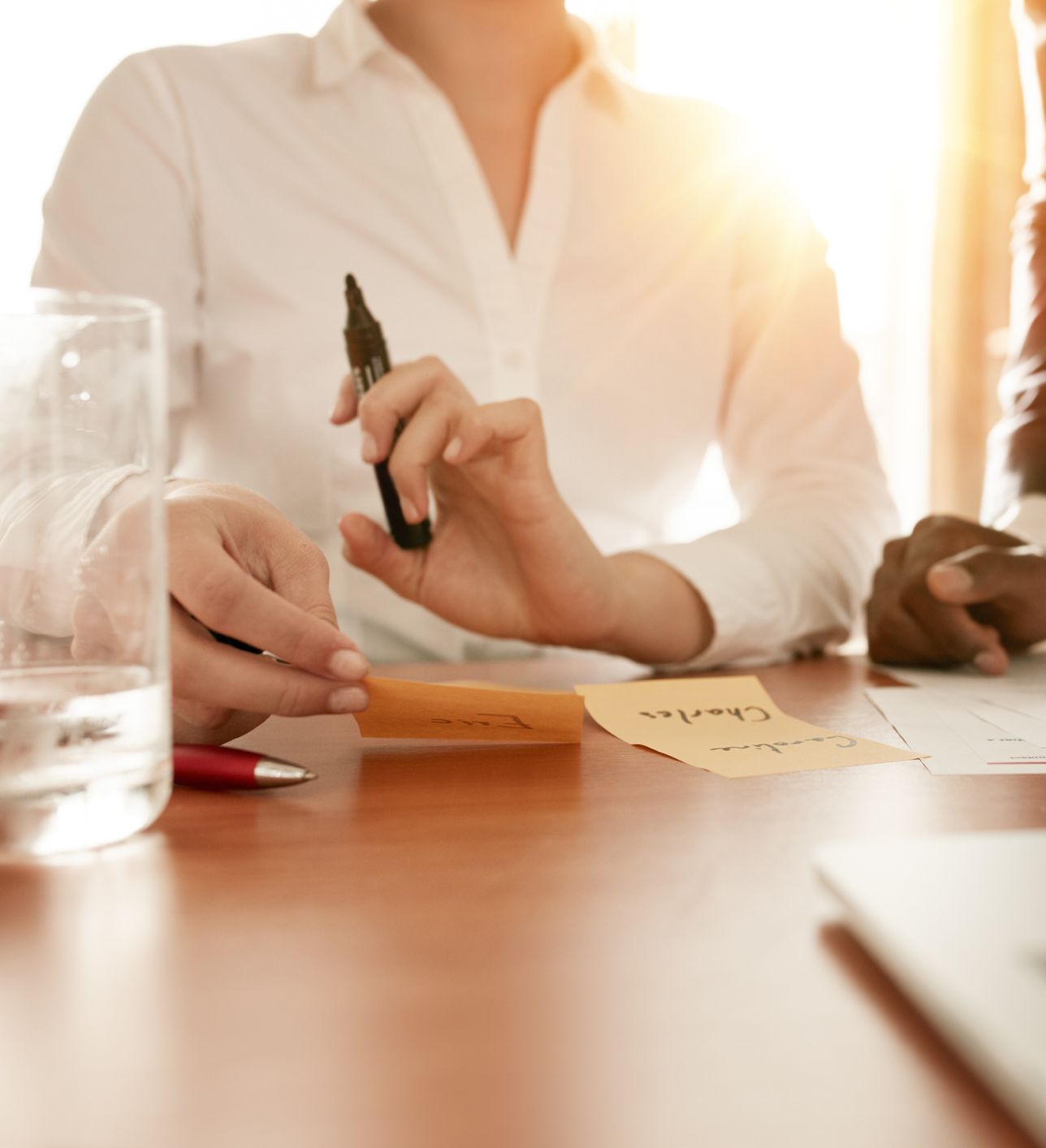 Two individuals in a business setting collaborate over documents and sticky notes on a table, with warm sunlight streaming in through a window. One person holds a marker.