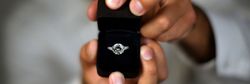 A close-up of hands holding an open black ring box with a sparkling diamond engagement ring, set against a blurred background.