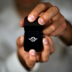 A close-up of hands holding an open black ring box with a sparkling diamond engagement ring, set against a blurred background.