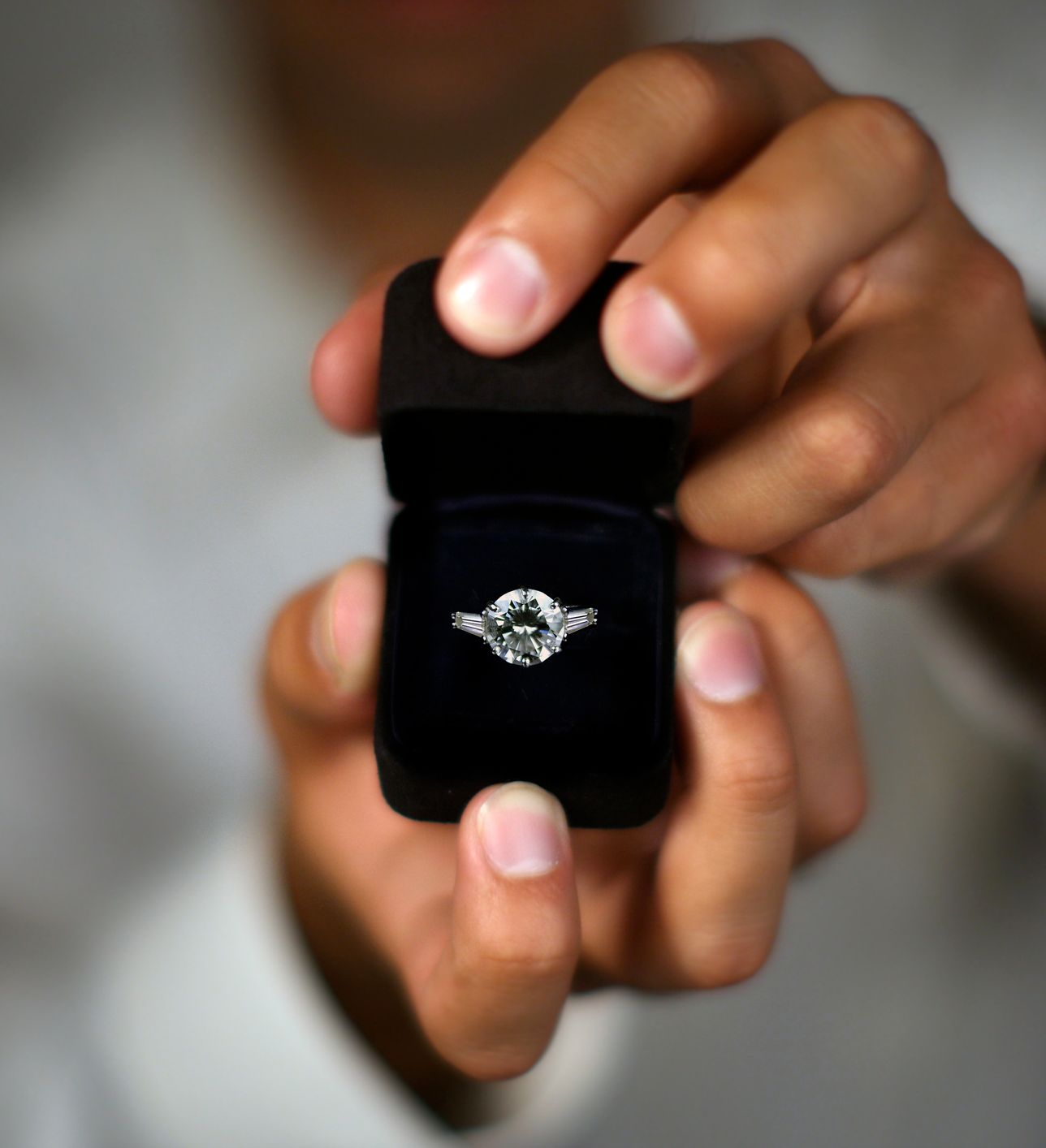 A close-up of hands holding an open black ring box with a sparkling diamond engagement ring, set against a blurred background.