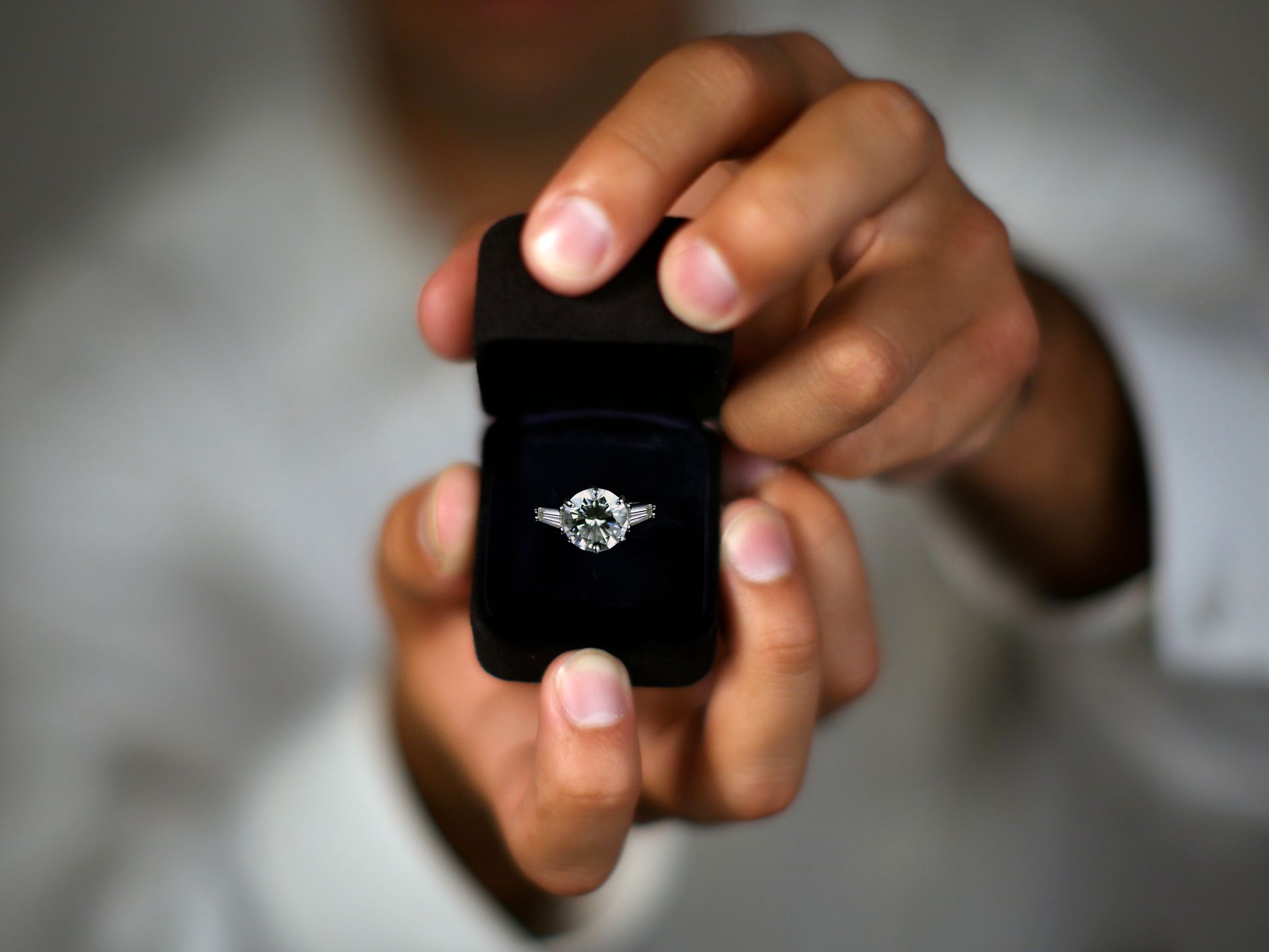 A close-up of hands holding an open black ring box with a sparkling diamond engagement ring, set against a blurred background.