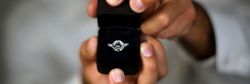 A close-up of hands holding an open black ring box with a sparkling diamond engagement ring, set against a blurred background.