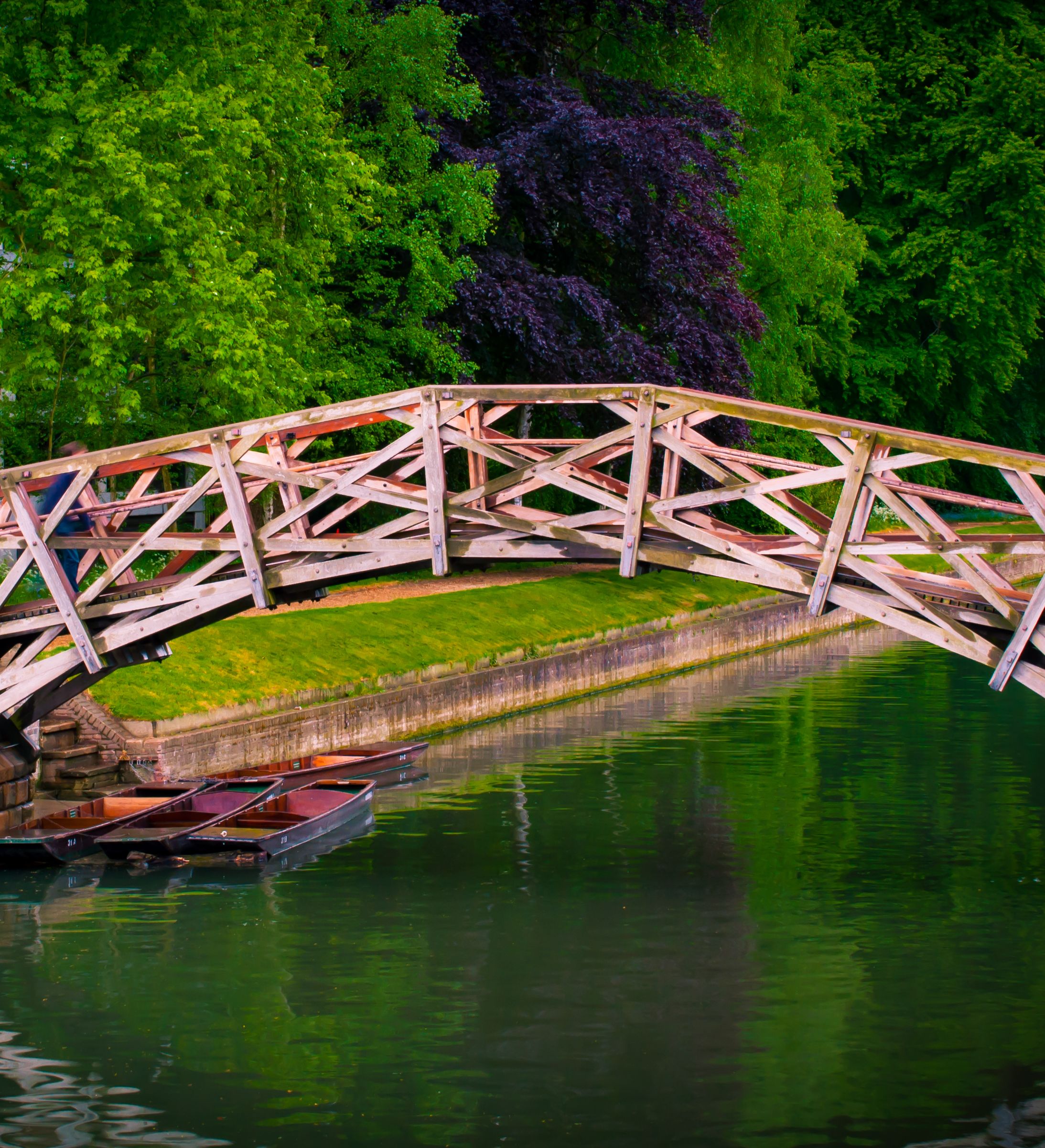 A wooden bridge with intricate crisscross patterns spans a calm river surrounded by lush green trees, with small boats docked beneath and a historic brick building nearby.