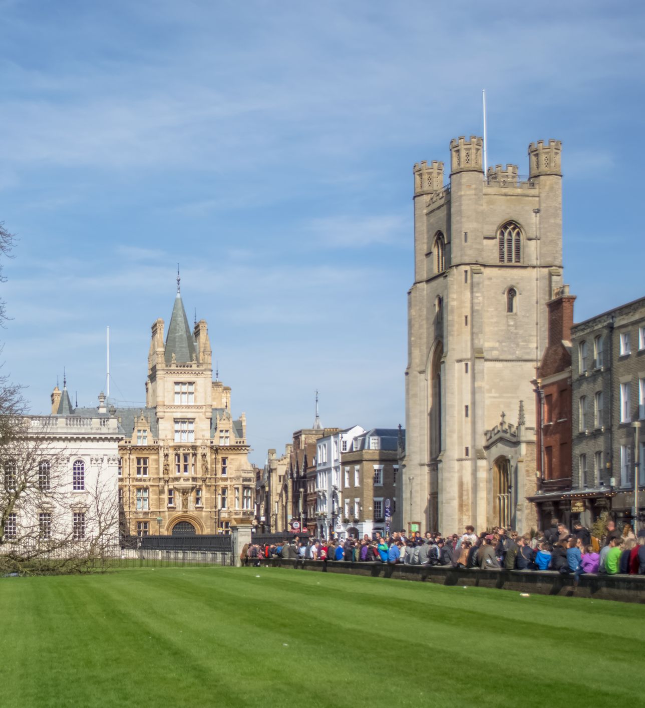 A historic cityscape featuring a large green lawn, a leafless tree, Gothic-style buildings, and a tall stone tower under a clear blue sky with people gathered along the path.