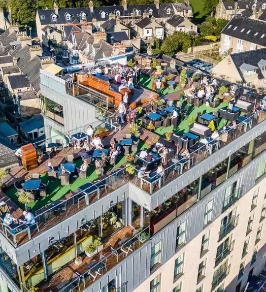 A vibrant rooftop bar and seating area atop a modern building, bustling with people enjoying drinks, surrounded by a cityscape of closely packed houses and streets below.