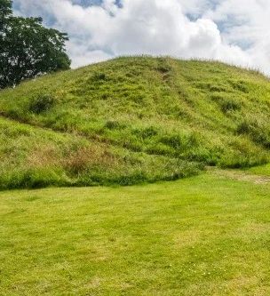 A grassy hill with visible walking paths winding up its slopes, surrounded by green lawn and trees, under a partly cloudy sky.