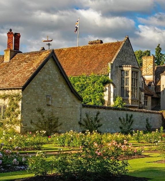 A historic stone manor with tall chimneys, surrounded by a vibrant rose garden in full bloom, neatly trimmed hedges, and lush green lawn under a partly cloudy sky.