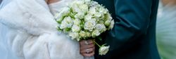 A close-up of a bride in a white fur wrap holding a bouquet of white roses with decorative accents, standing next to a groom in a dark suit.