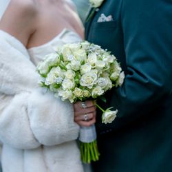 A close-up of a bride in a white fur wrap holding a bouquet of white roses with decorative accents, standing next to a groom in a dark suit.