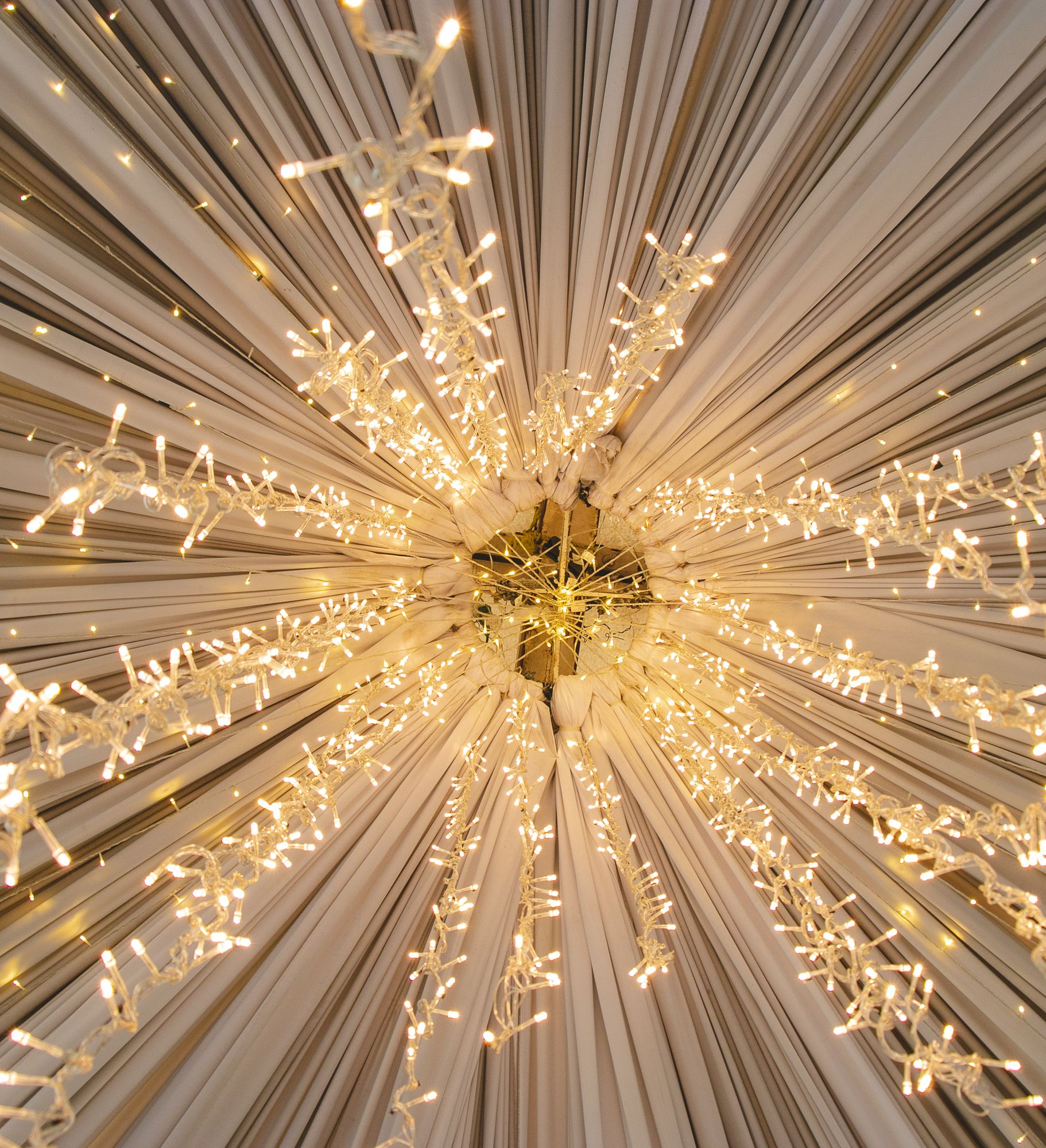 A ceiling decorated with white drapes arranged in a radial pattern and illuminated by strings of warm white fairy lights radiating outward from a central point.