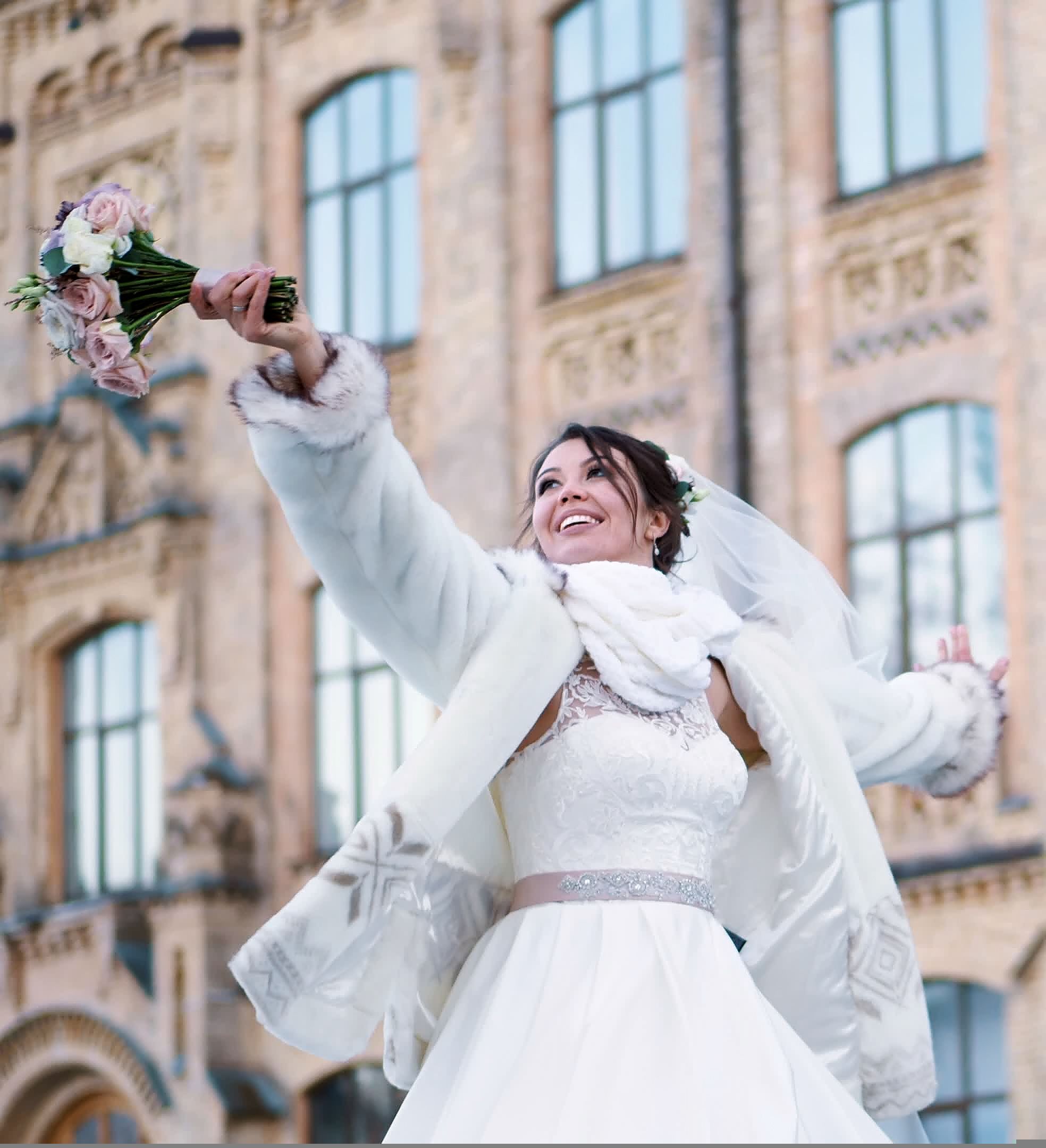 A joyful bride in a white gown and fur wrap holding a bouquet aloft, standing outside a historic building with ornate brickwork and large arched windows.