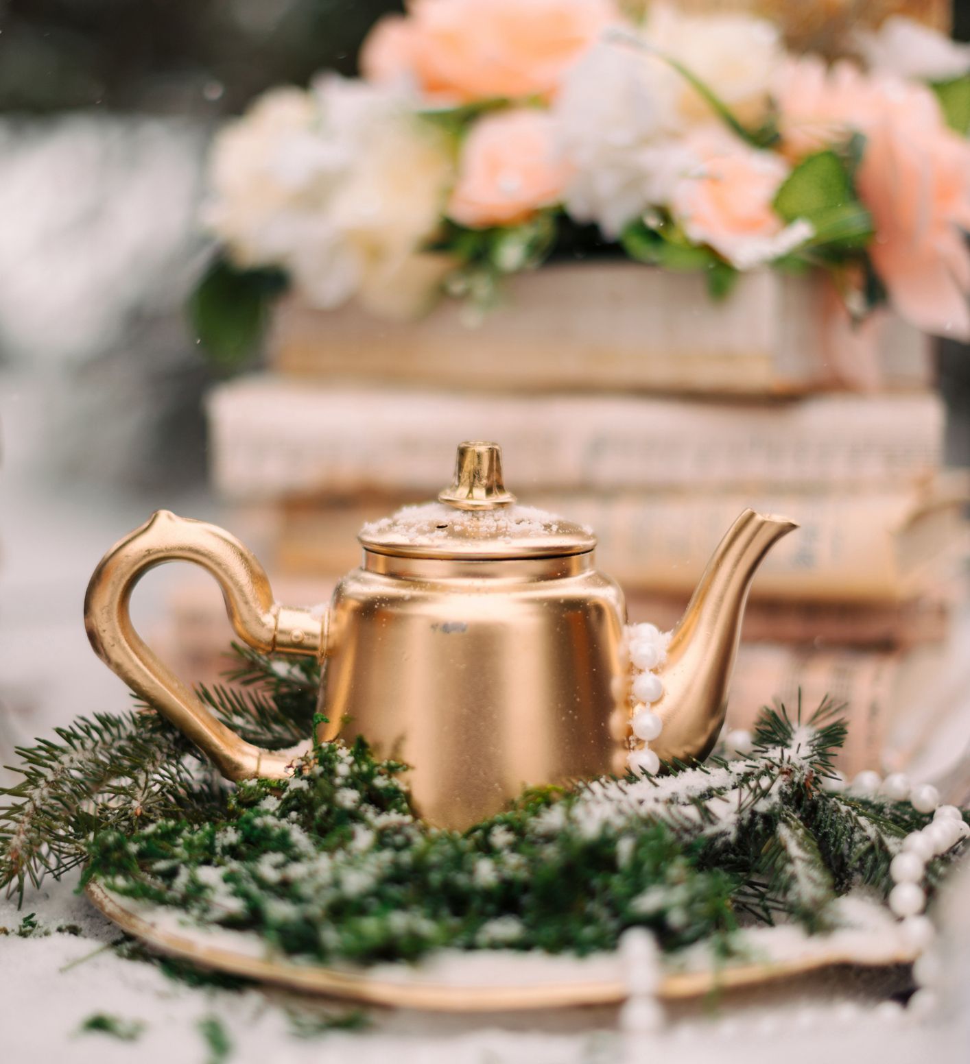 A vintage gold teapot adorned with snow and pearls, placed on a bed of evergreen branches, with blurred books and pastel flowers in the background.