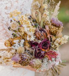 A close-up of a bride holding a rustic bouquet of dried flowers, including roses, thistles, and grasses, with a lace wedding gown visible in the background.