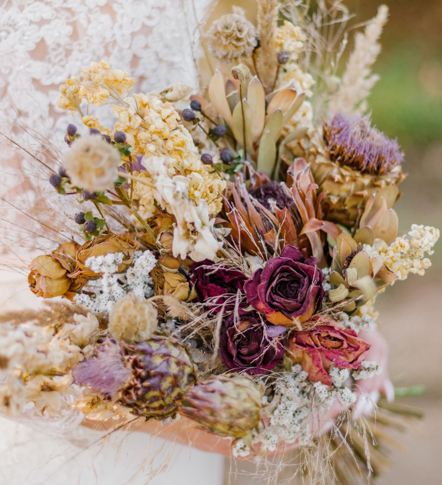 A close-up of a bride holding a rustic bouquet of dried flowers, including roses, thistles, and grasses, with a lace wedding gown visible in the background.