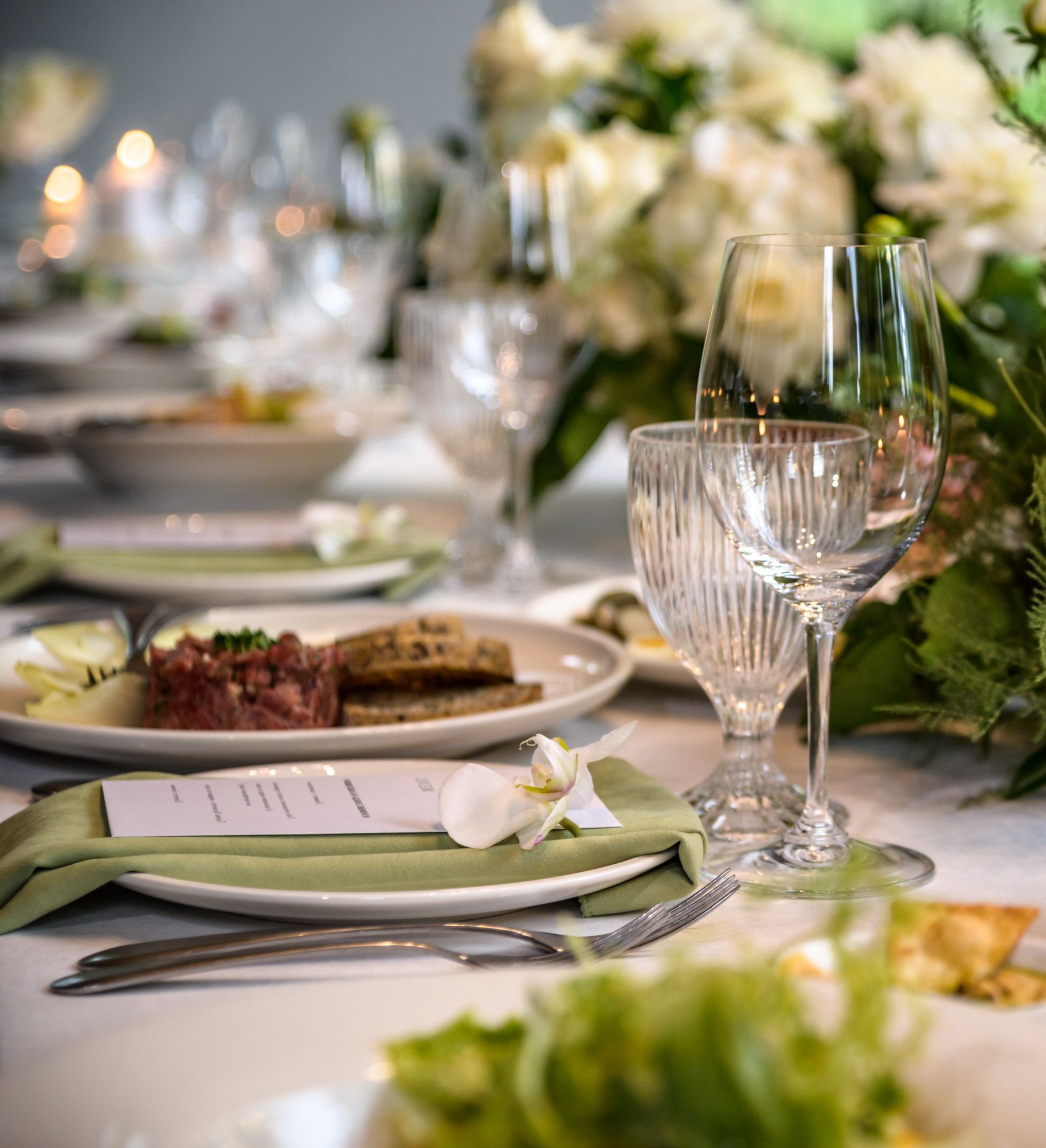 A beautifully set dining table with elegant glassware, green napkins, white plates, lit candles, and lush floral arrangements featuring white and green accents.