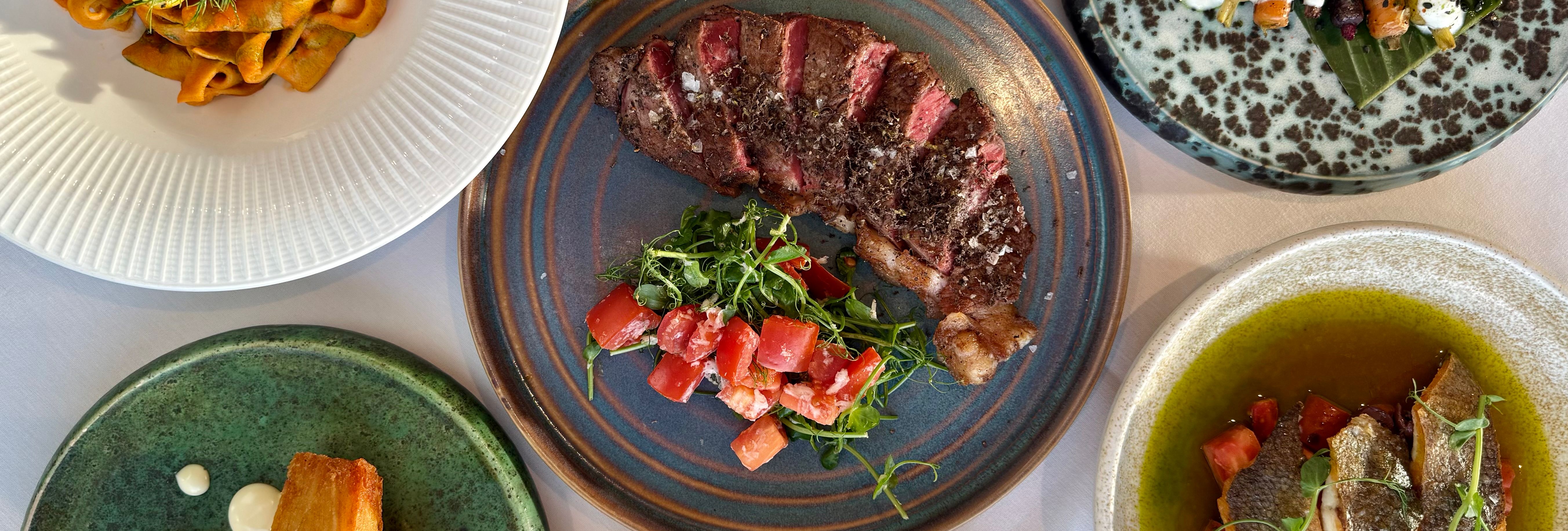 A top-down view of a gourmet meal with various plated dishes, including steak, pasta with shrimp, crispy potatoes, fish, and colorful vegetable sides.
