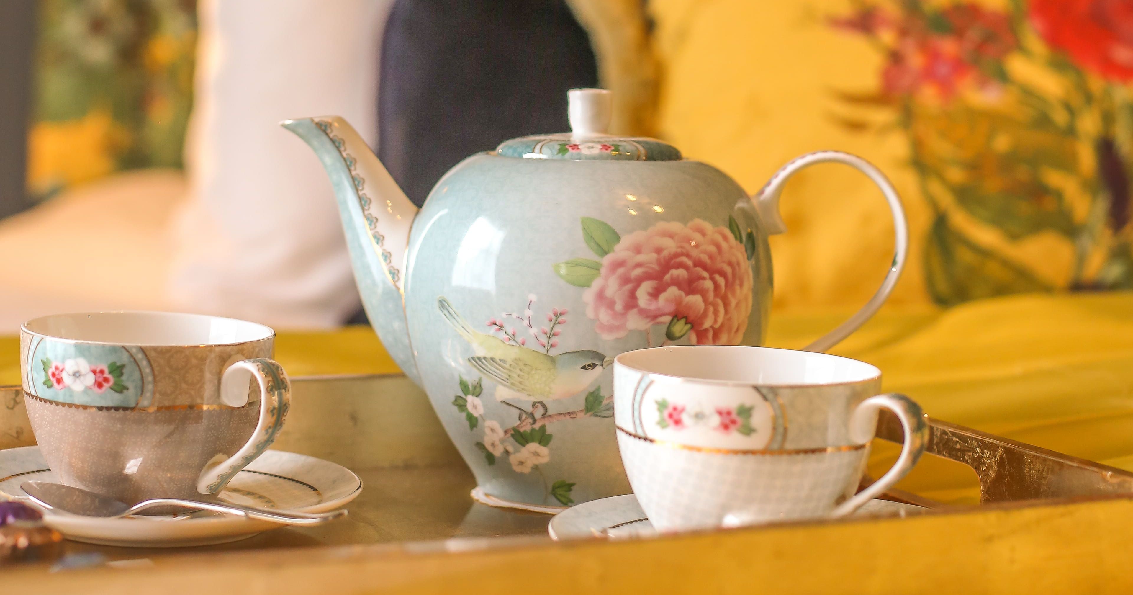 A floral teapot and two teacups on a tray atop the bed in the Rosa feature room at Gonville Hotel, with vibrant cushions and a floral headboard.