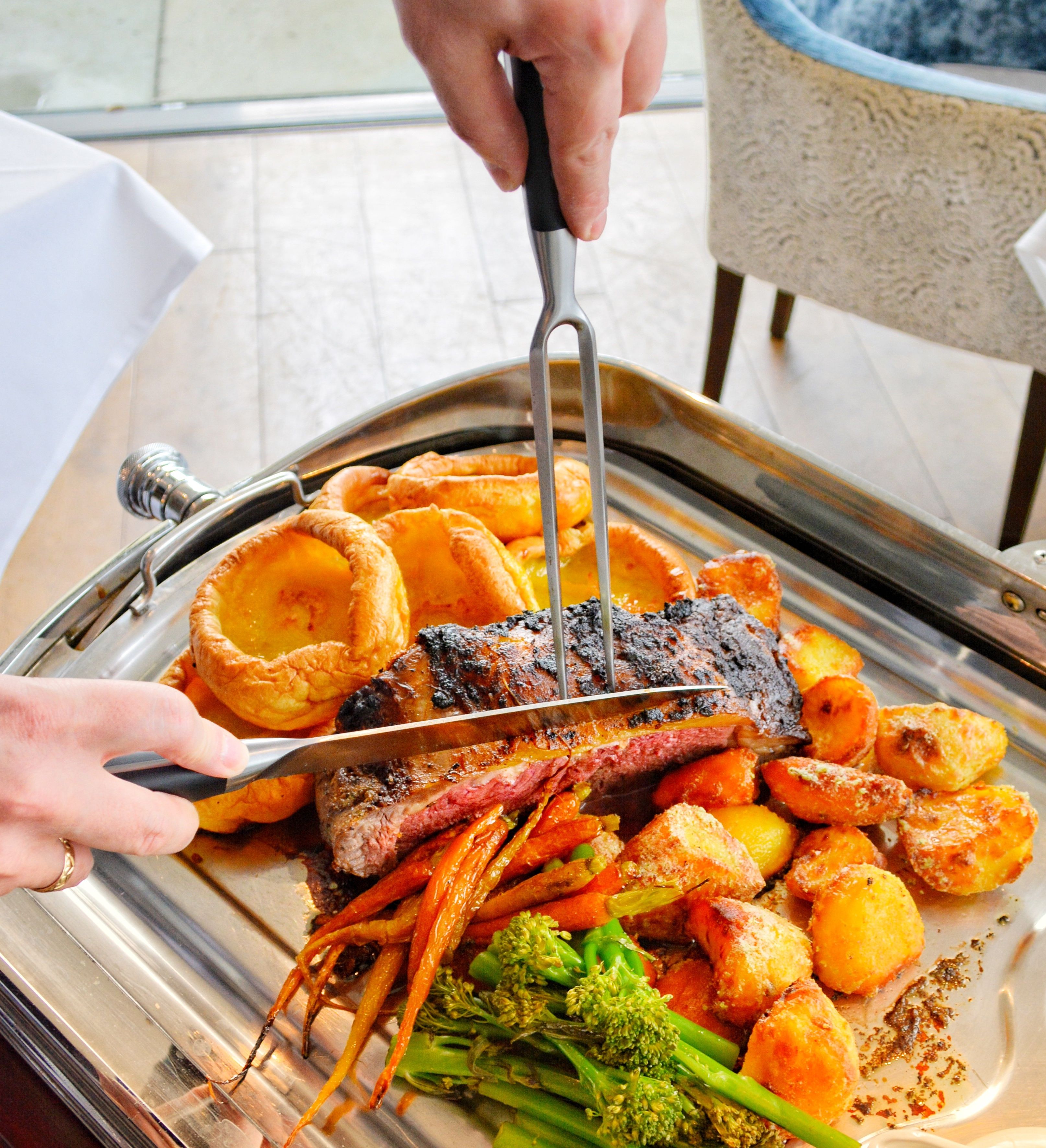 A person carves a medium-rare roast beef on a silver tray, surrounded by golden roasted potatoes, Yorkshire puddings, glazed carrots, and green broccolini.