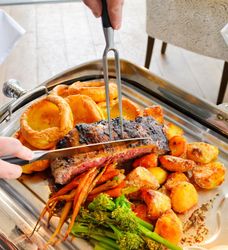A person carves a medium-rare roast beef on a silver tray, surrounded by golden roasted potatoes, Yorkshire puddings, glazed carrots, and green broccolini.