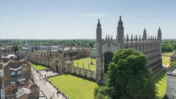 Aerial view of King's College Chapel and its surrounding courtyard in Cambridge, with tourists walking along nearby paths on a bright, sunny day.