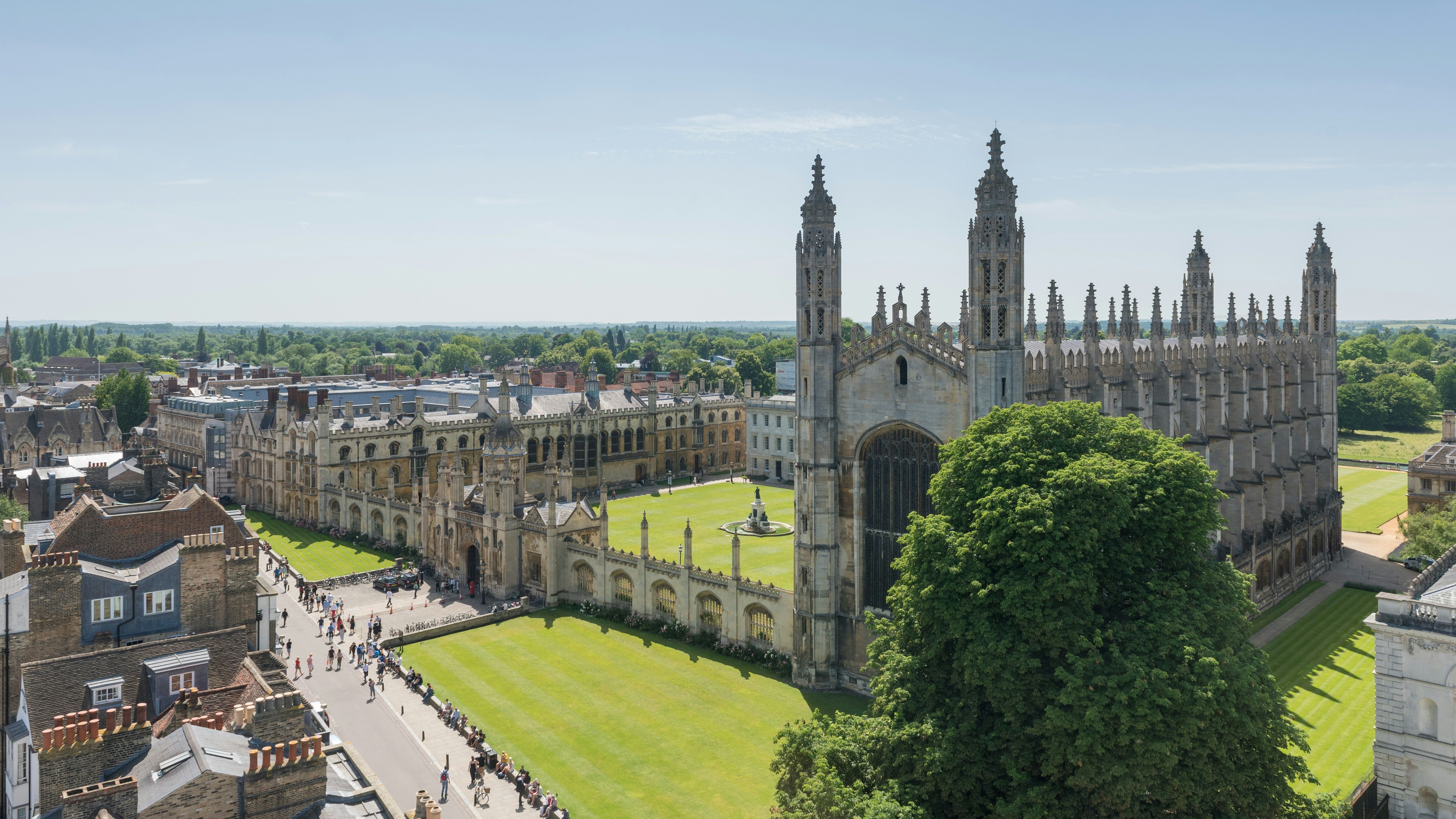 Aerial view of King's College Chapel and its surrounding courtyard in Cambridge, with tourists walking along nearby paths on a bright, sunny day.