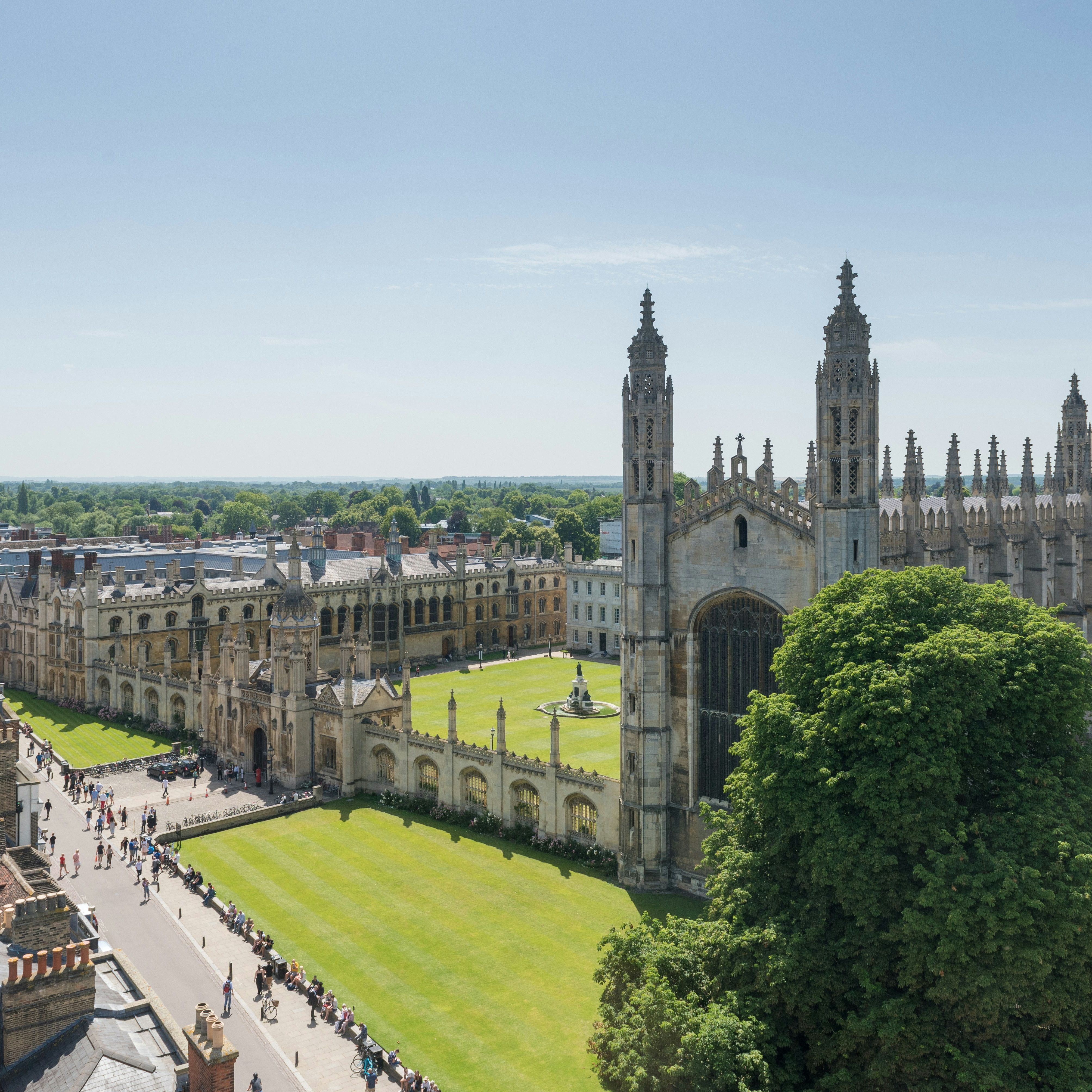 Aerial view of King's College Chapel and its surrounding courtyard in Cambridge, with tourists walking along nearby paths on a bright, sunny day.