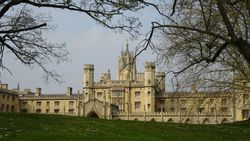 Historic Gothic-style college building with tall towers and arched windows, framed by leafless trees and a grassy lawn under a pale blue sky.