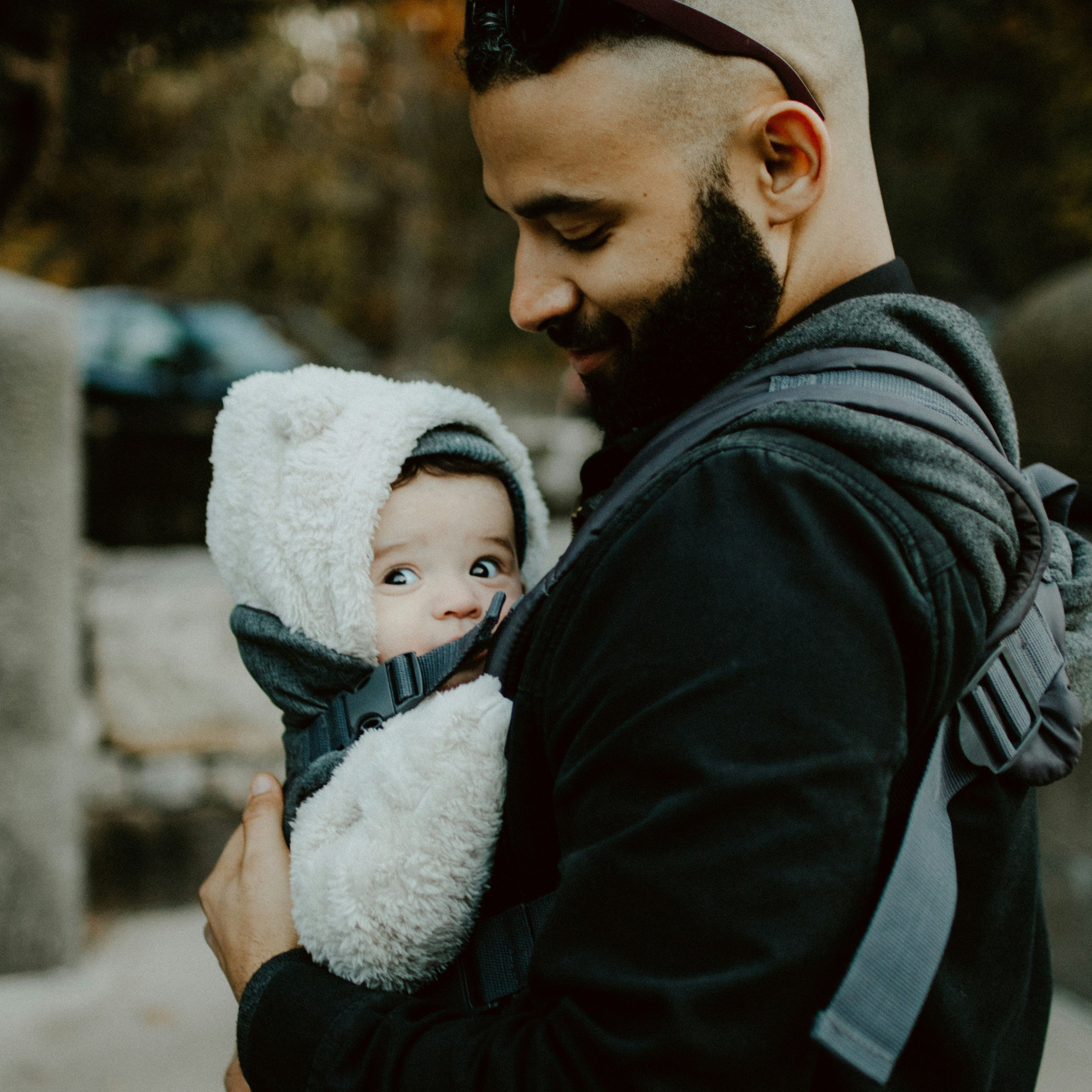 Smiling man with a beard carrying a bundled-up baby in a front carrier on a chilly day; the baby looks to the side while snuggled in a fluffy hooded outfit.