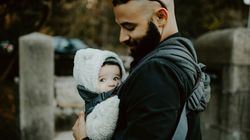 Smiling man with a beard carrying a bundled-up baby in a front carrier on a chilly day; the baby looks to the side while snuggled in a fluffy hooded outfit.