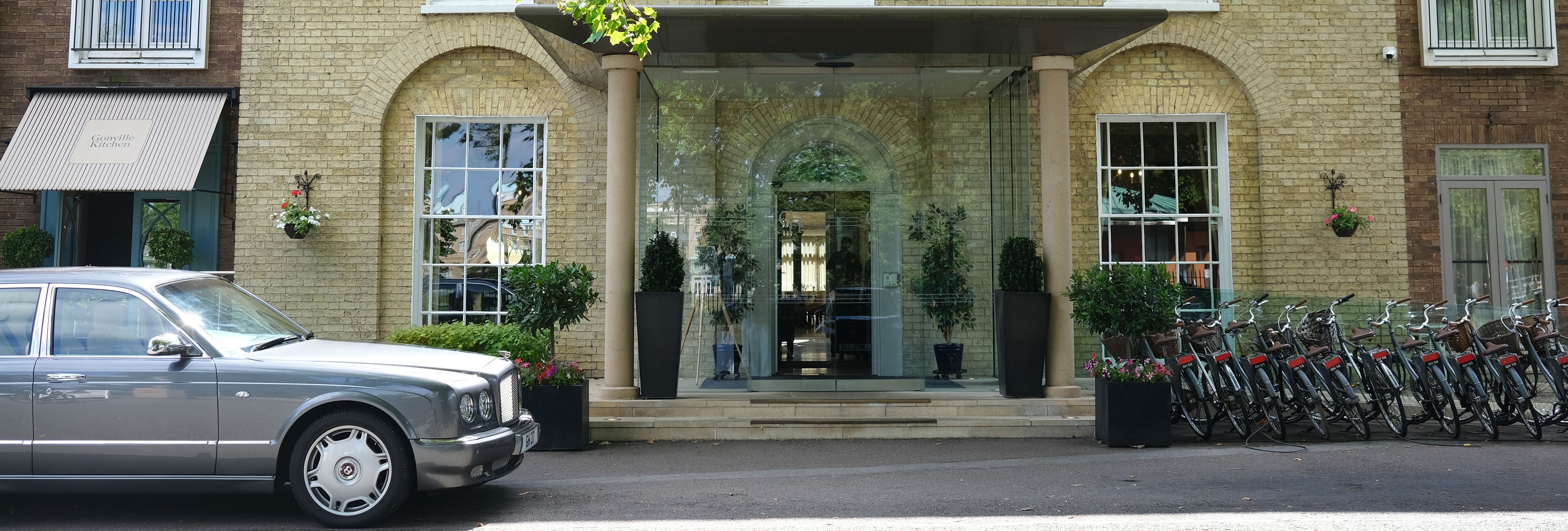 Hotel entrance with glass doors, potted plants, parked classic car, and row of bicycles