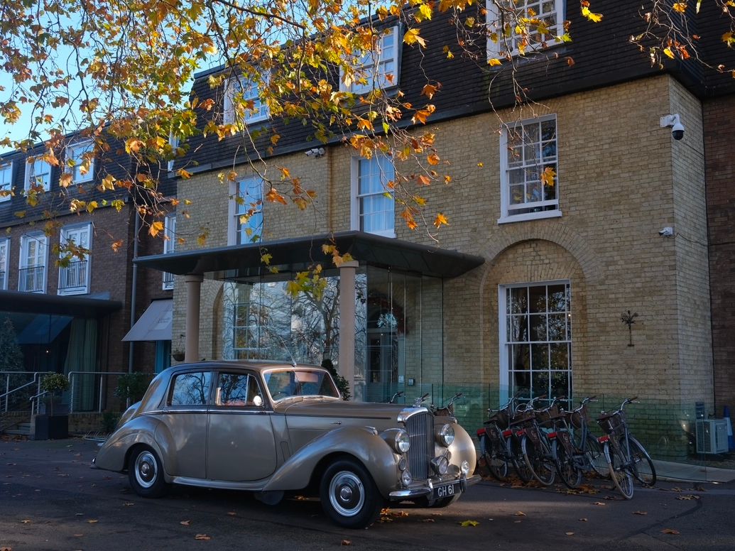 Classic silver car parked in front of a brick building with autumn leaves overhead