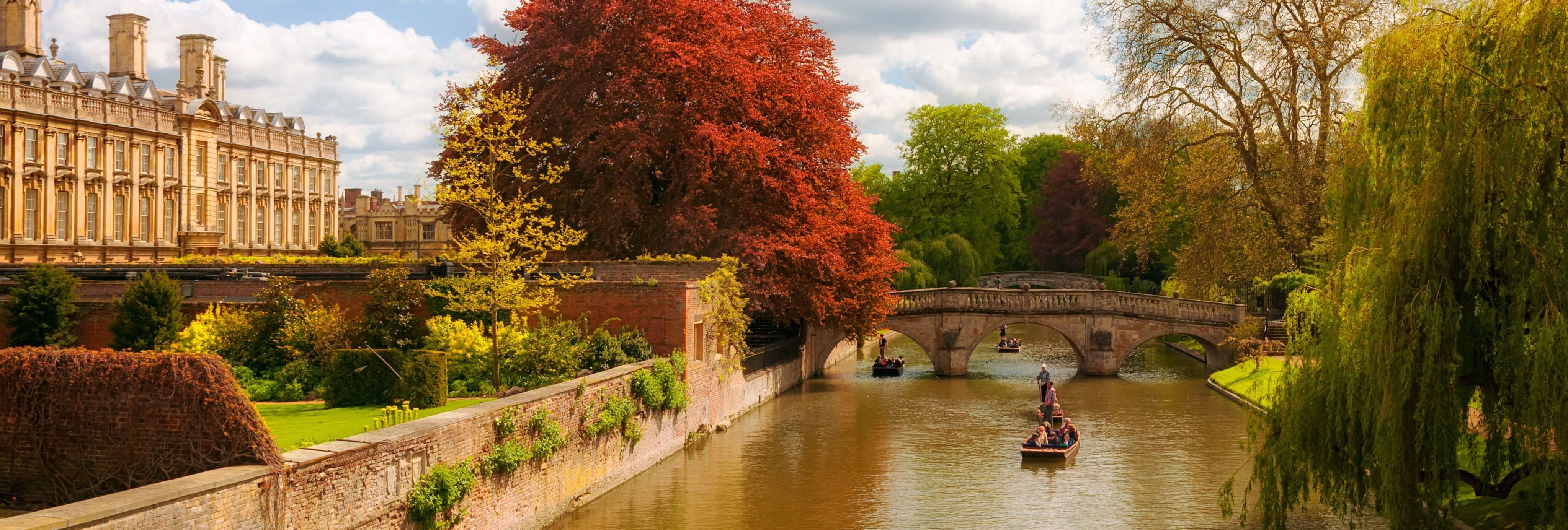Autumn scene in Cambridge with people punting on the river, trees with colorful foliage, and historic college buildings.