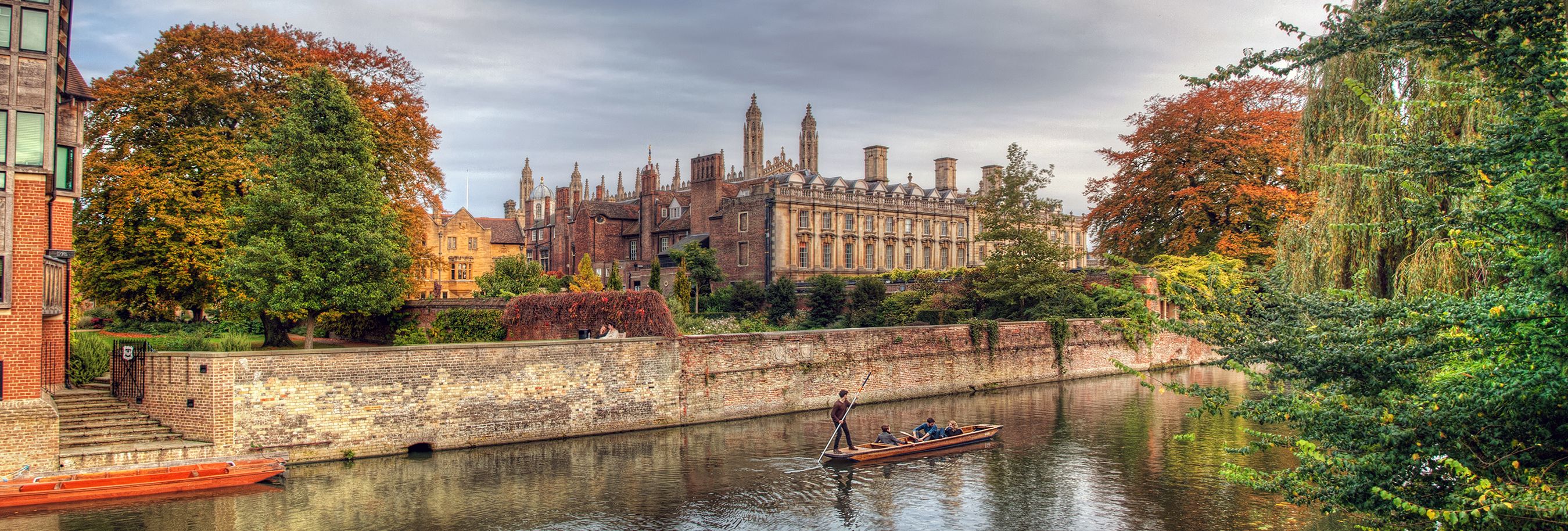 A scenic autumn view of Cambridge with colorful trees, historic university buildings, and people punting on the river.