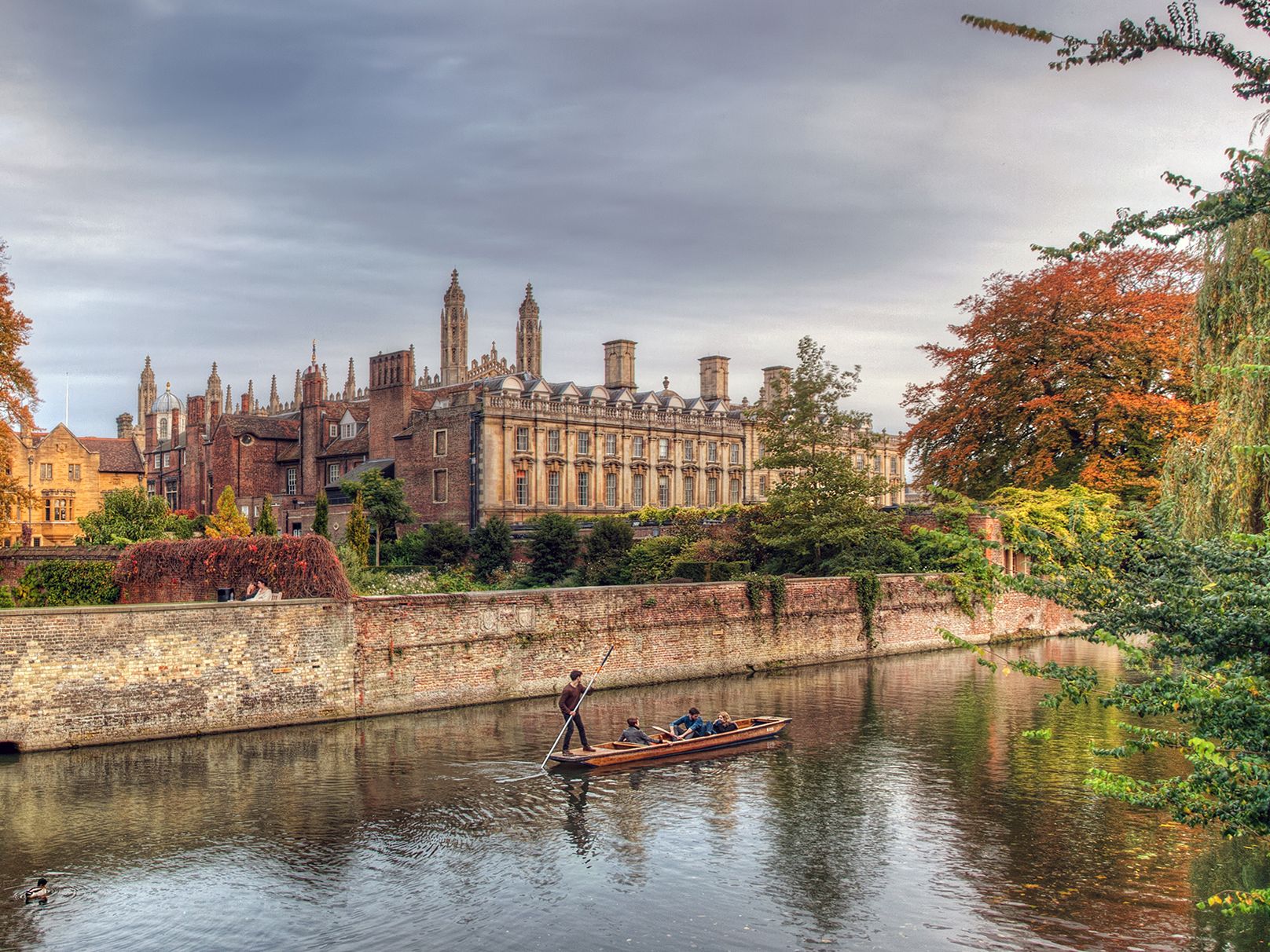 A scenic autumn view of Cambridge with colorful trees, historic university buildings, and people punting on the river.
