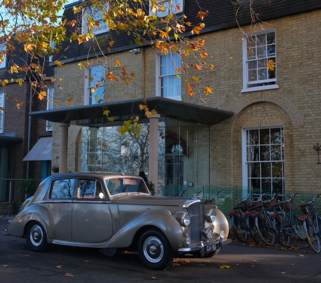 Classic vintage car parked in front of a building with autumn leaves on tree branches overhead.