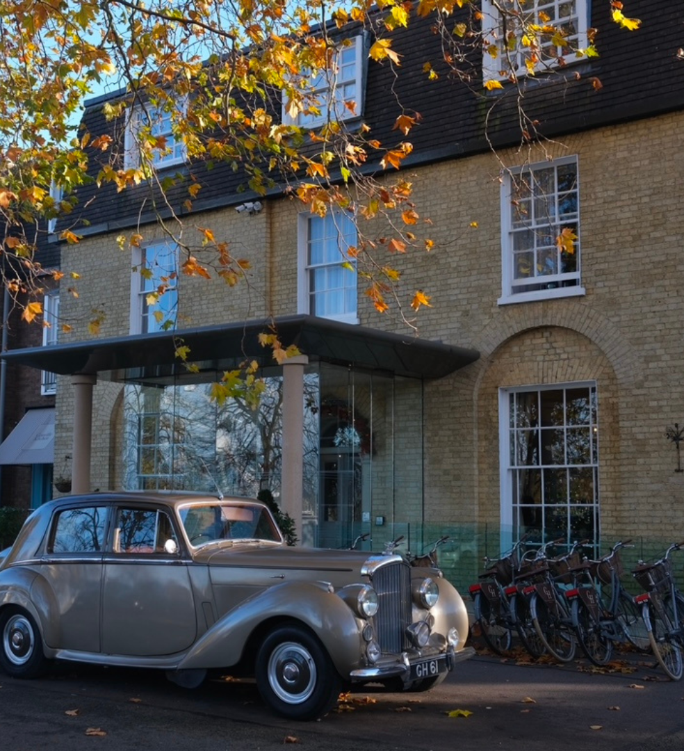 Classic vintage car parked in front of a building with autumn leaves on tree branches overhead.