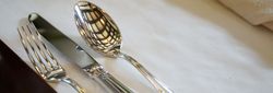 A fork, knife, and spoon with ornate handles neatly placed on a white tablecloth, with a napkin nearby and a ceiling reflection on the spoon.