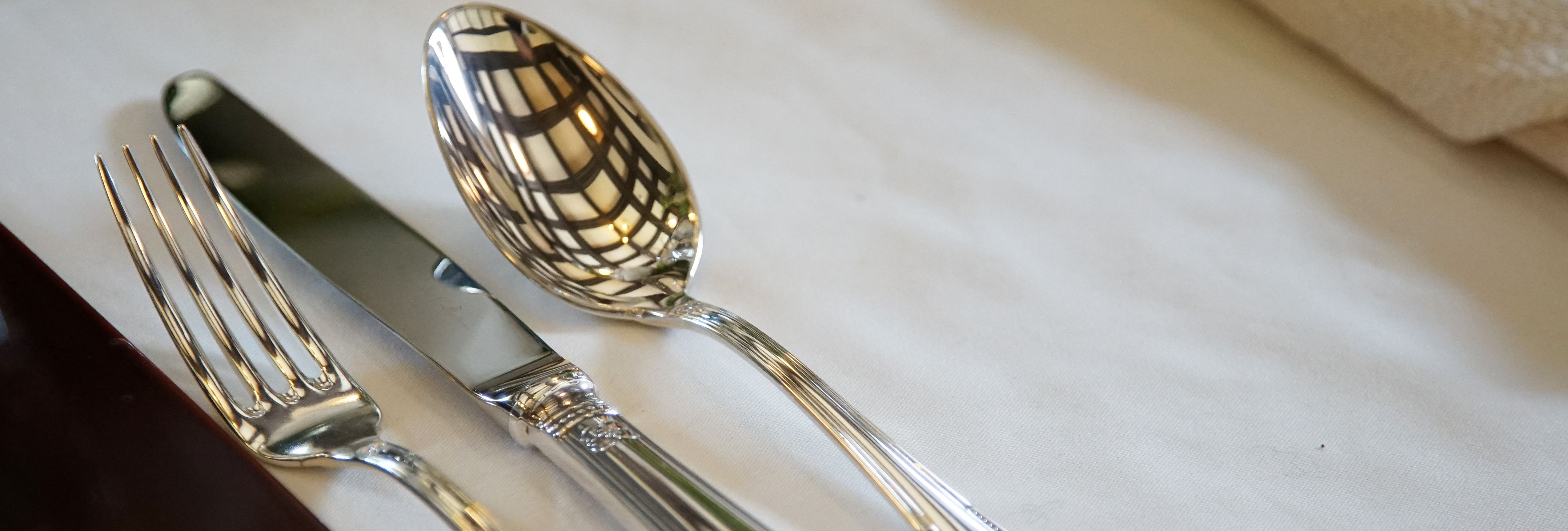 A fork, knife, and spoon with ornate handles neatly placed on a white tablecloth, with a napkin nearby and a ceiling reflection on the spoon.