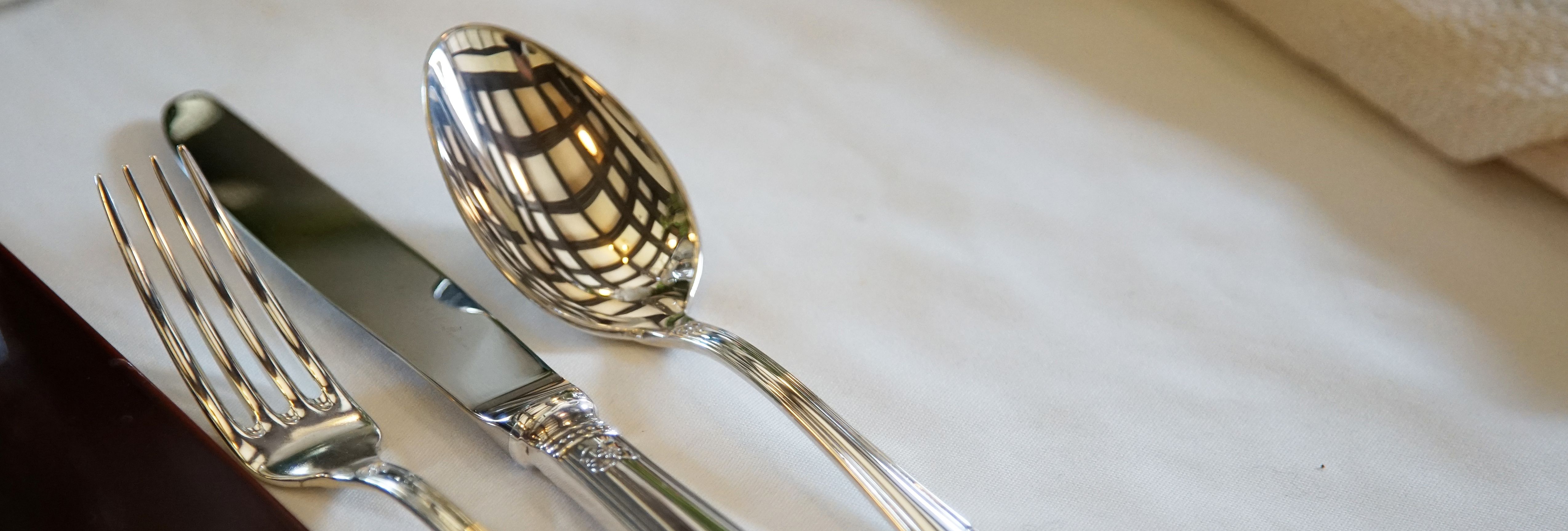 A fork, knife, and spoon with ornate handles neatly placed on a white tablecloth, with a napkin nearby and a ceiling reflection on the spoon.