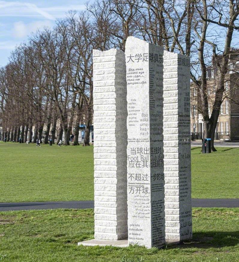 Tall stone sculpture engraved with text in multiple languages, standing on a grassy area outdoors.