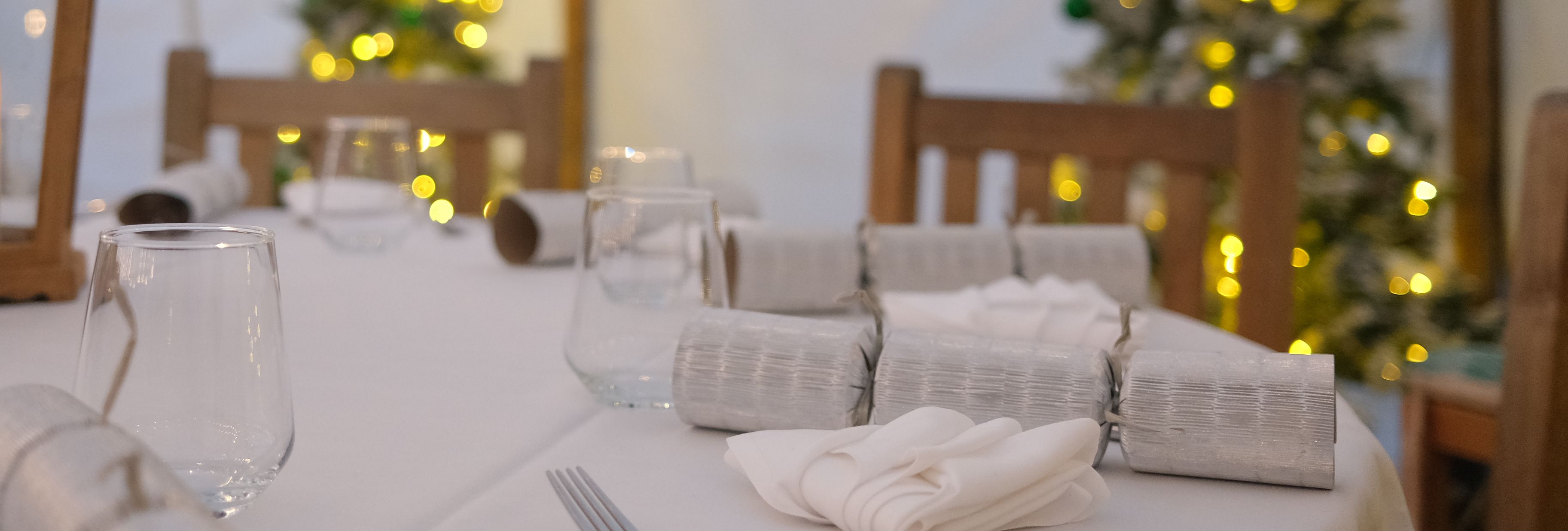 Festive dining table set with crackers, lantern, and glassware, with decorated Christmas trees in the background