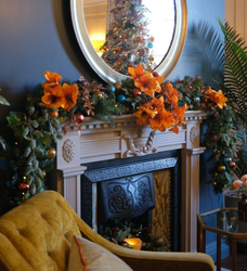 A decorated fireplace with orange flowers, greenery, and ornaments, with a Christmas tree reflected in a round mirror above.