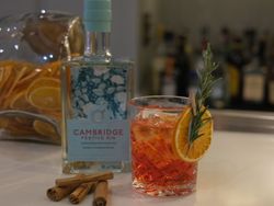 A bottle of Cambridge Festive Gin with a cocktail garnished with orange slice and rosemary, cinnamon sticks, and jars of dried fruit on a bar counter.