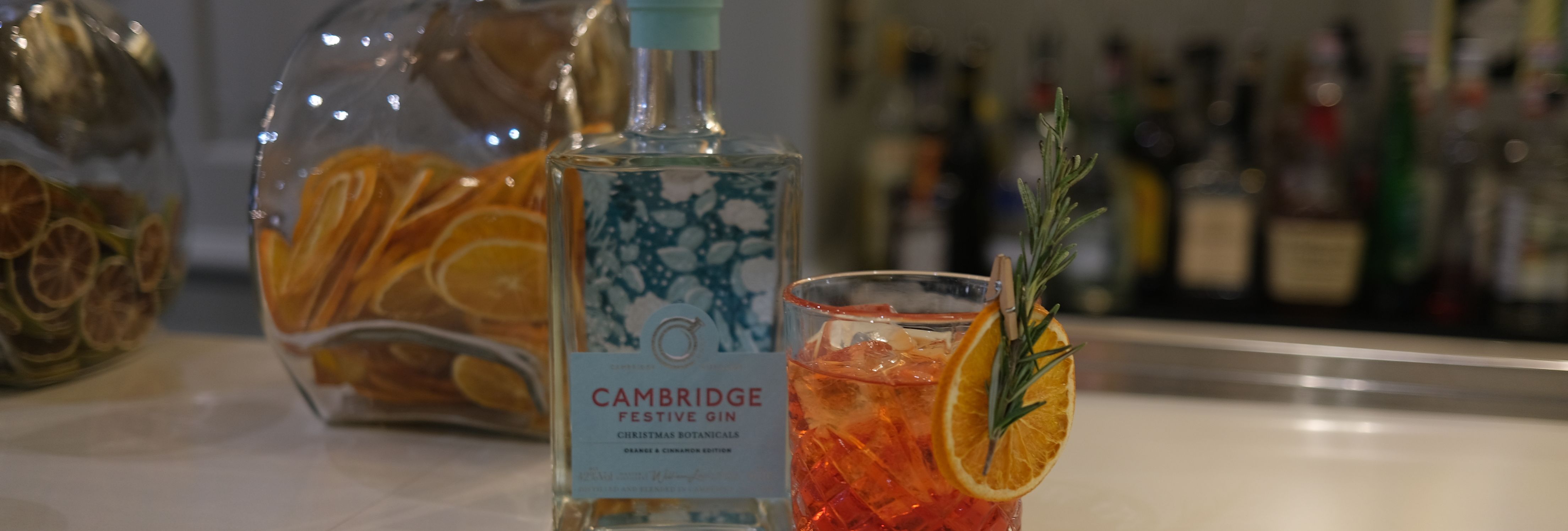 A bottle of Cambridge Festive Gin with a cocktail garnished with orange slice and rosemary, cinnamon sticks, and jars of dried fruit on a bar counter.