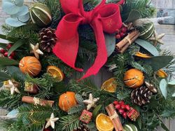 Festive Christmas wreath decorated with a large red bow, dried oranges, pinecones, cinnamon sticks, small presents, and greenery.