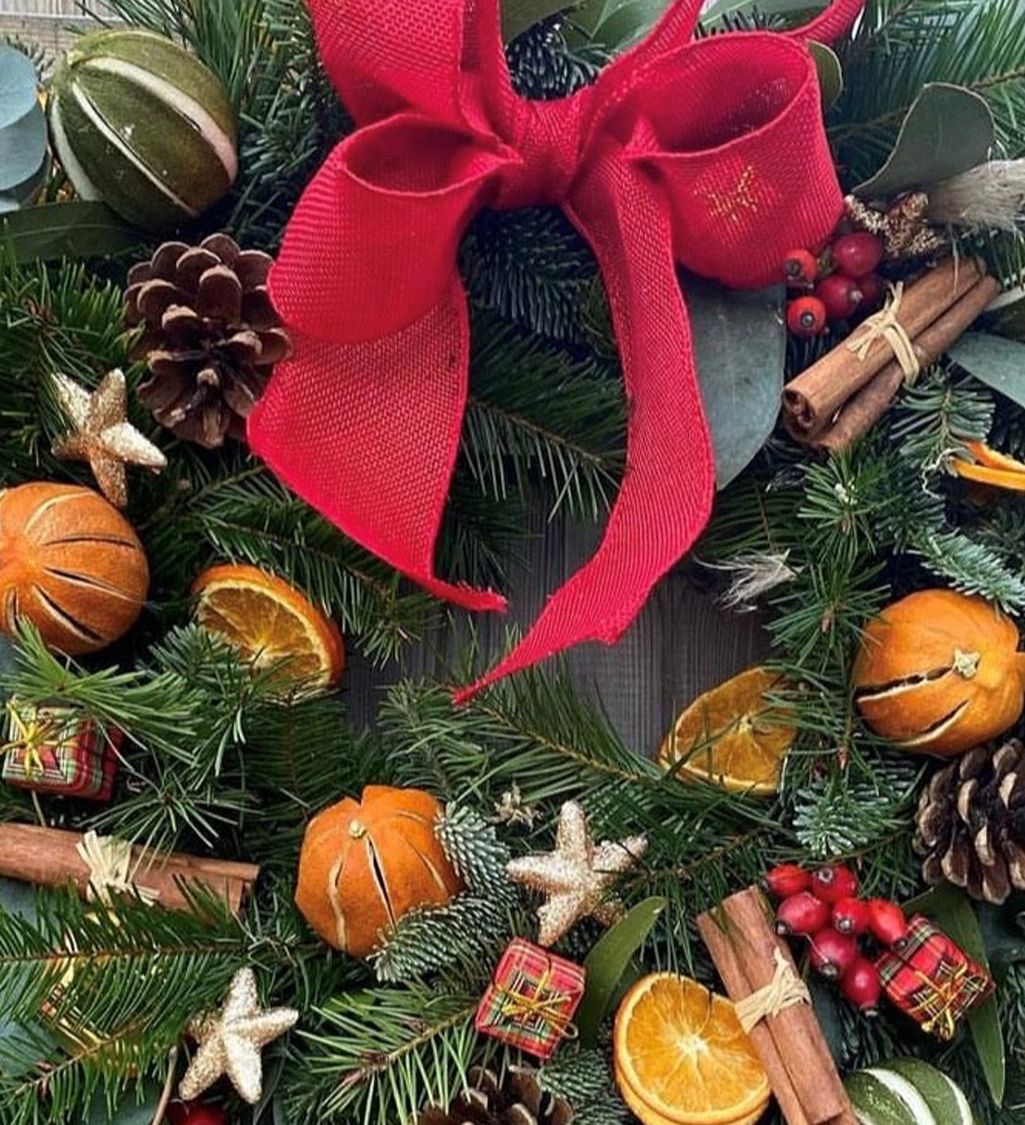 Festive Christmas wreath decorated with a large red bow, dried oranges, pinecones, cinnamon sticks, small presents, and greenery.