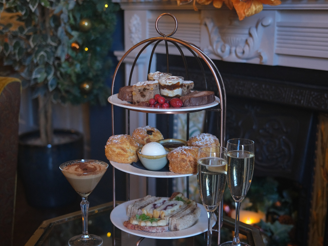 Three-tiered afternoon tea stand with assorted pastries, scones, and sandwiches, accompanied by two glasses of champagne and a cocktail on a glass table.