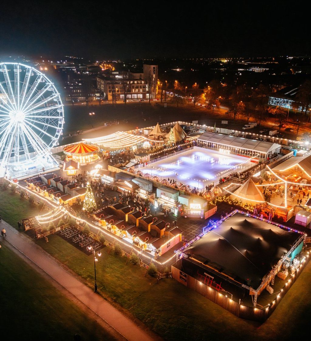 A brightly lit carnival at night with a large Ferris wheel, ice skating rink, and festive market booths